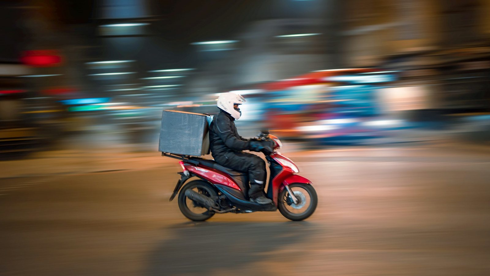 Entregador de scooter vermelha com baú de entrega numa rua da cidade à noite